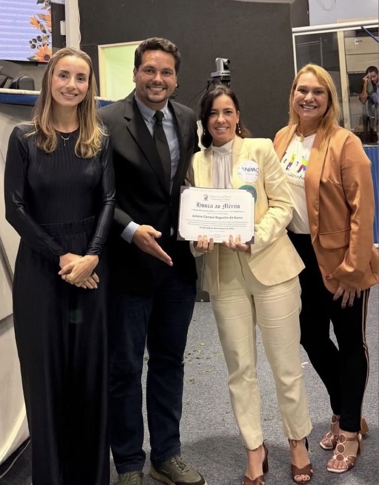 Juliana Gama segura o certificado de Honra ao Mérito ao lado de três pessoas no plenário da Câmara